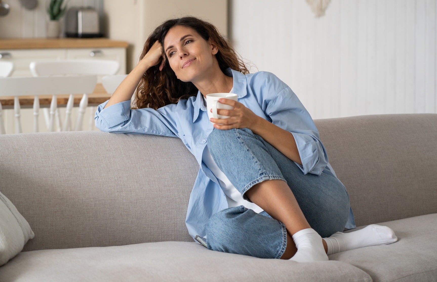 Young happy relaxed woman sitting on sofa enjoying quiet moments at home with cup of tea or coffee